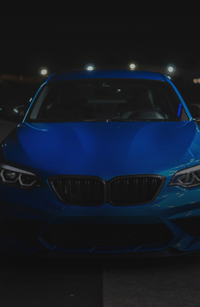 Blue BMW M2 sports car parked at night, illuminated by headlights and overhead lights - Bimmerfix