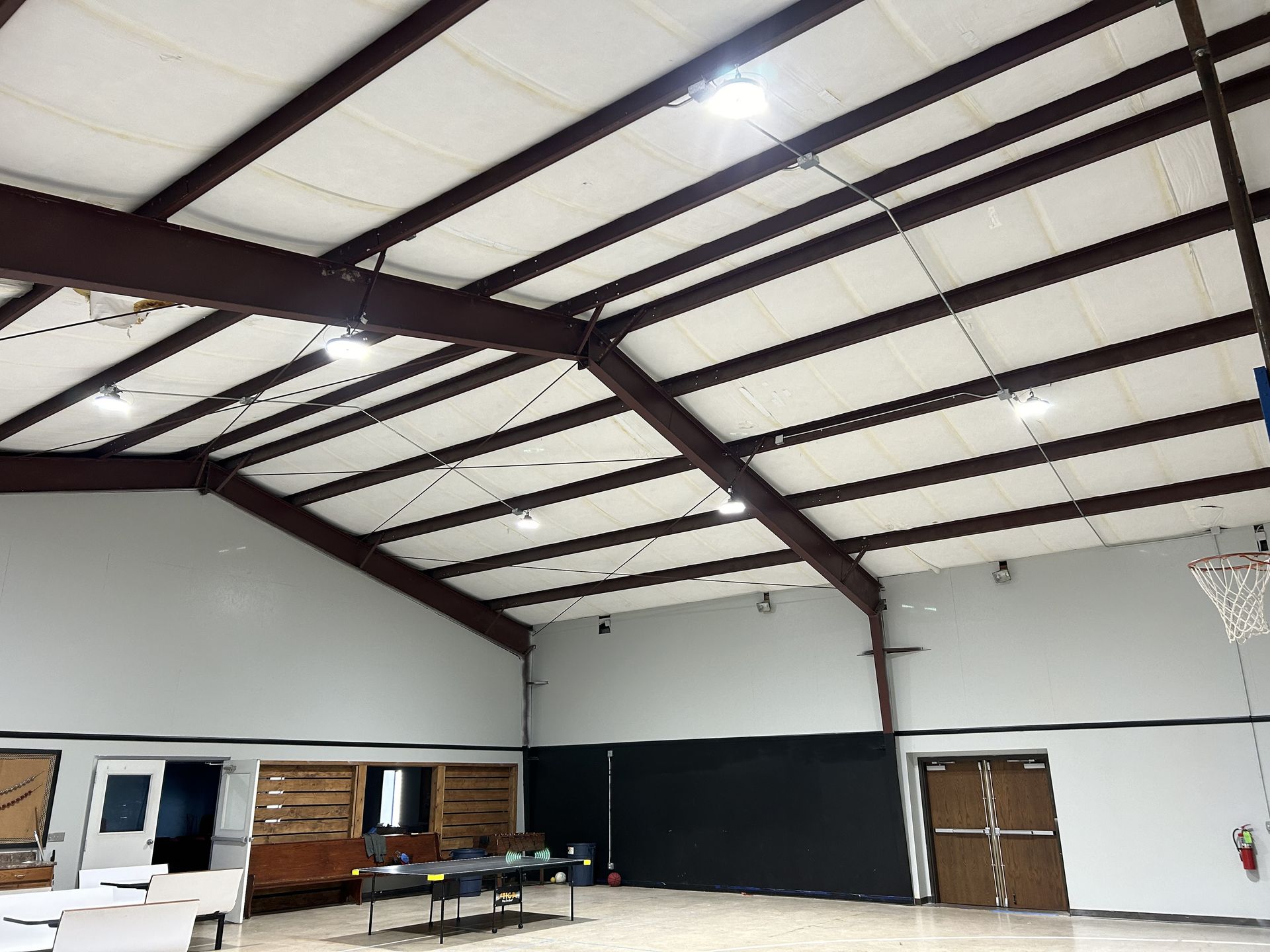 Interior view of a gymnasium with a high ceiling, metal beams, and bright lights. Walls are white and dark.