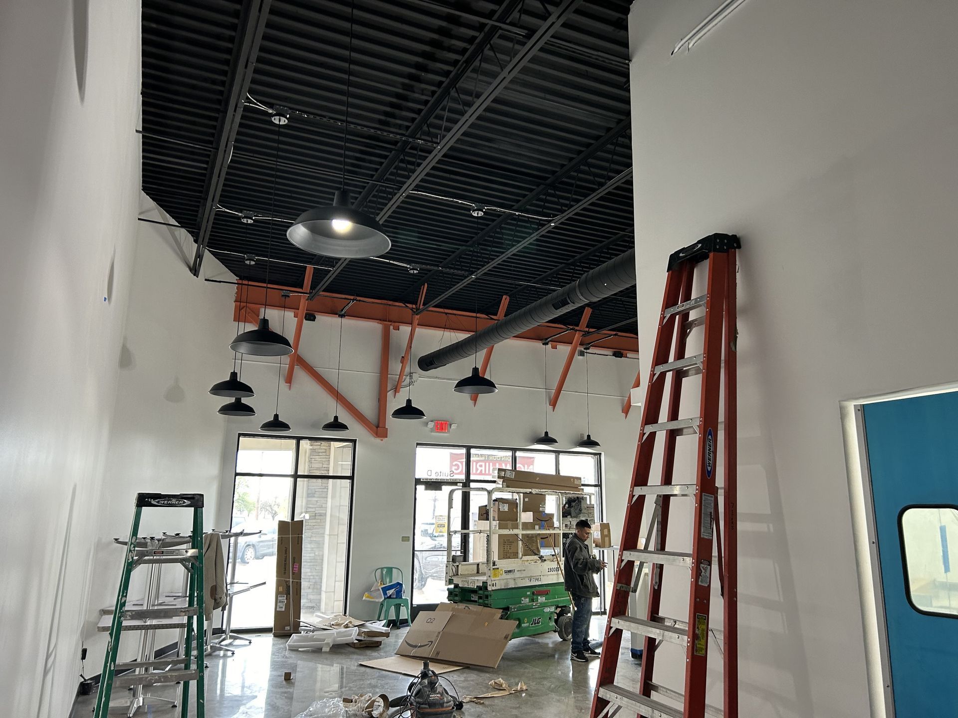 Interior construction site. Worker near a ladder; exposed ceiling with lights; large windows and a door.