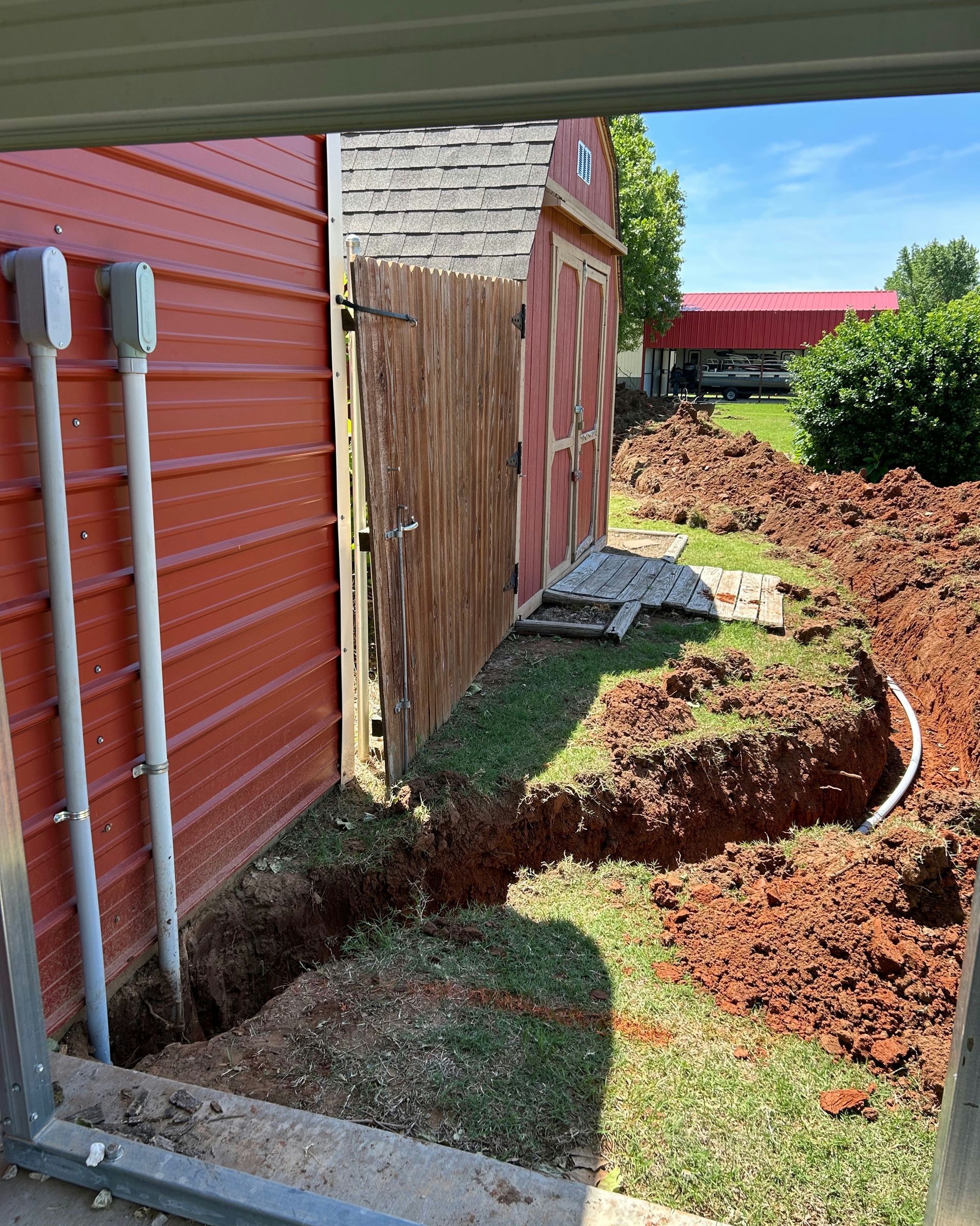 Exterior view of a red building with conduit and trenches dug along the side, leading to a wooden shed.