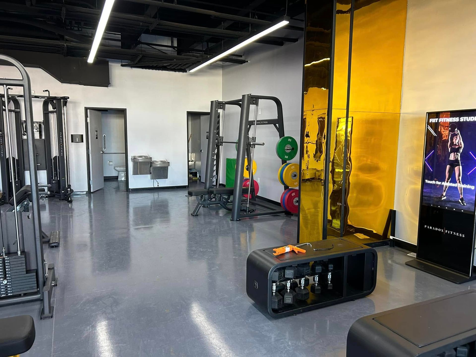 Gym interior with exercise equipment, including weight rack and benches, with a mirrored yellow accent wall.