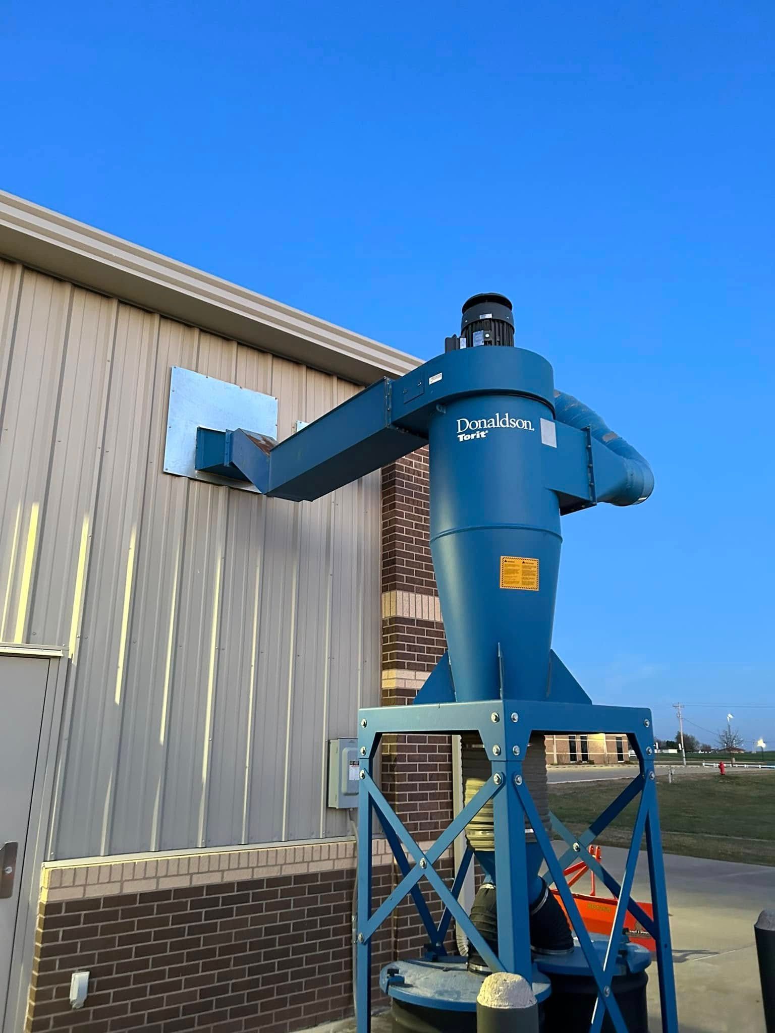 Blue industrial dust collector mounted on a metal frame, exterior wall with blue sky.