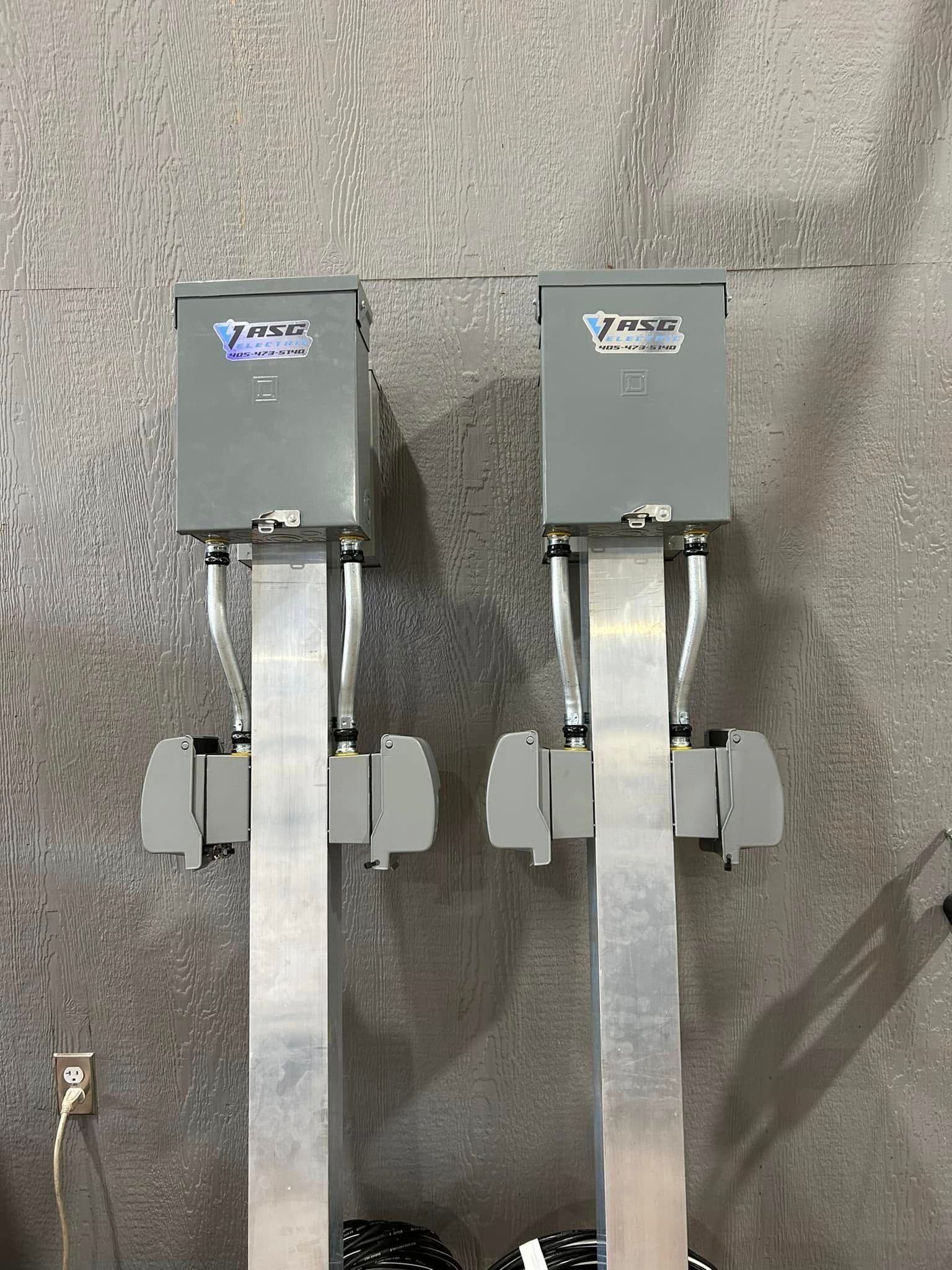 Two gray electrical boxes mounted on silver metal poles, against a mottled gray wall.