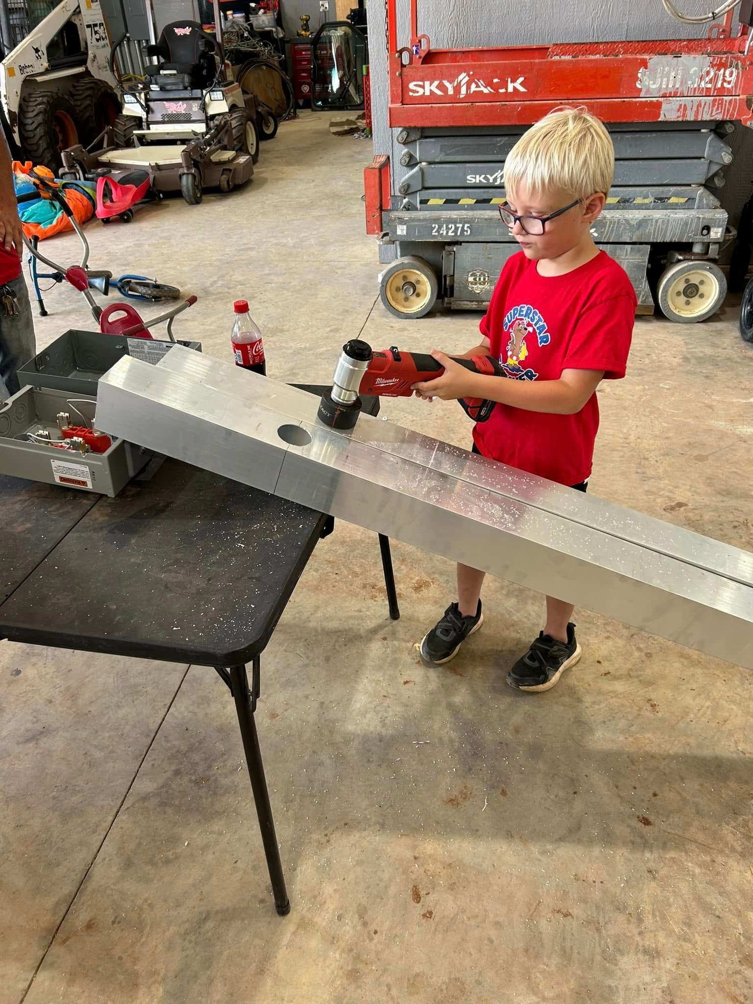 Boy in red shirt using a grinder on metal, standing near a table in a workshop.