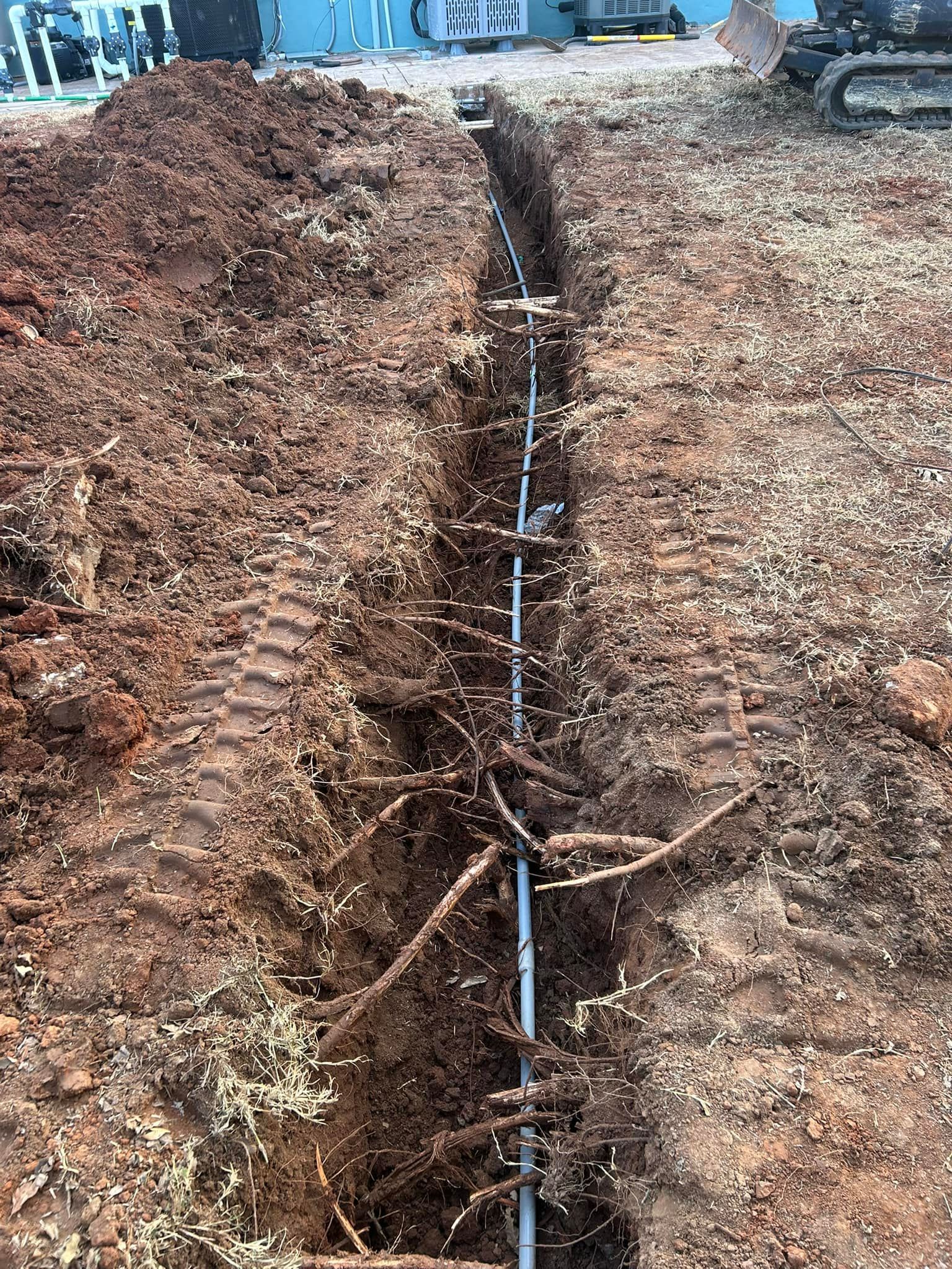 A trench with a gray pipe running through it, surrounded by dirt and tree roots.