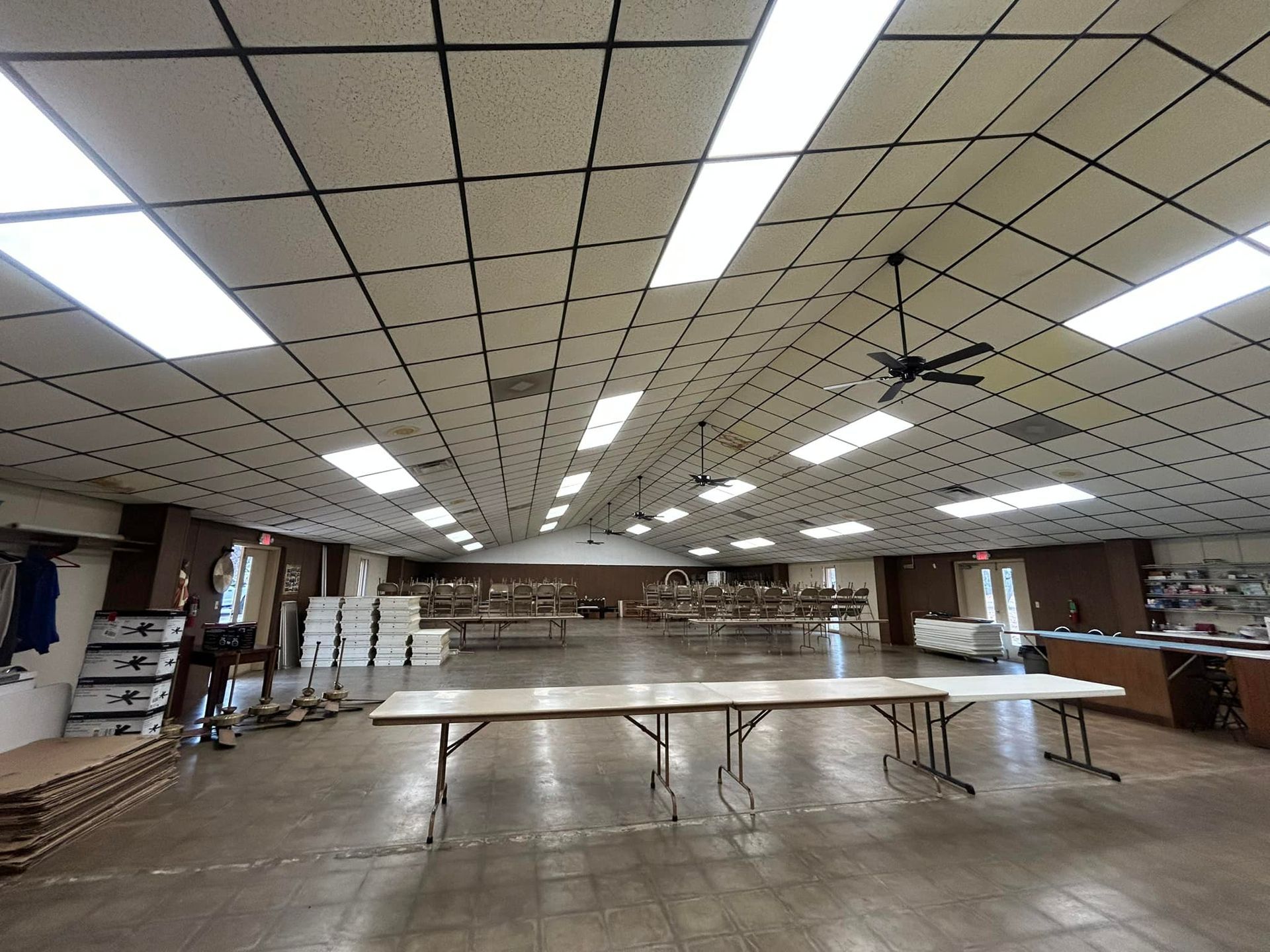 Large empty hall with folding tables set up. Ceiling grid with fluorescent lights, ceiling fan.