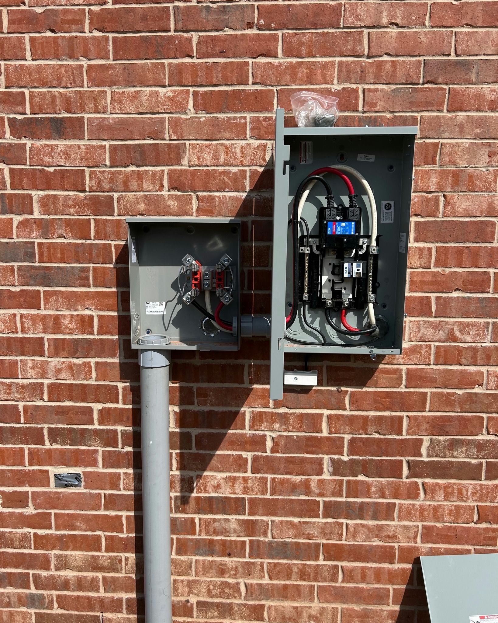 Exterior electrical panel with open cover mounted on brick wall. Gray metal, wires, and conduits are visible.