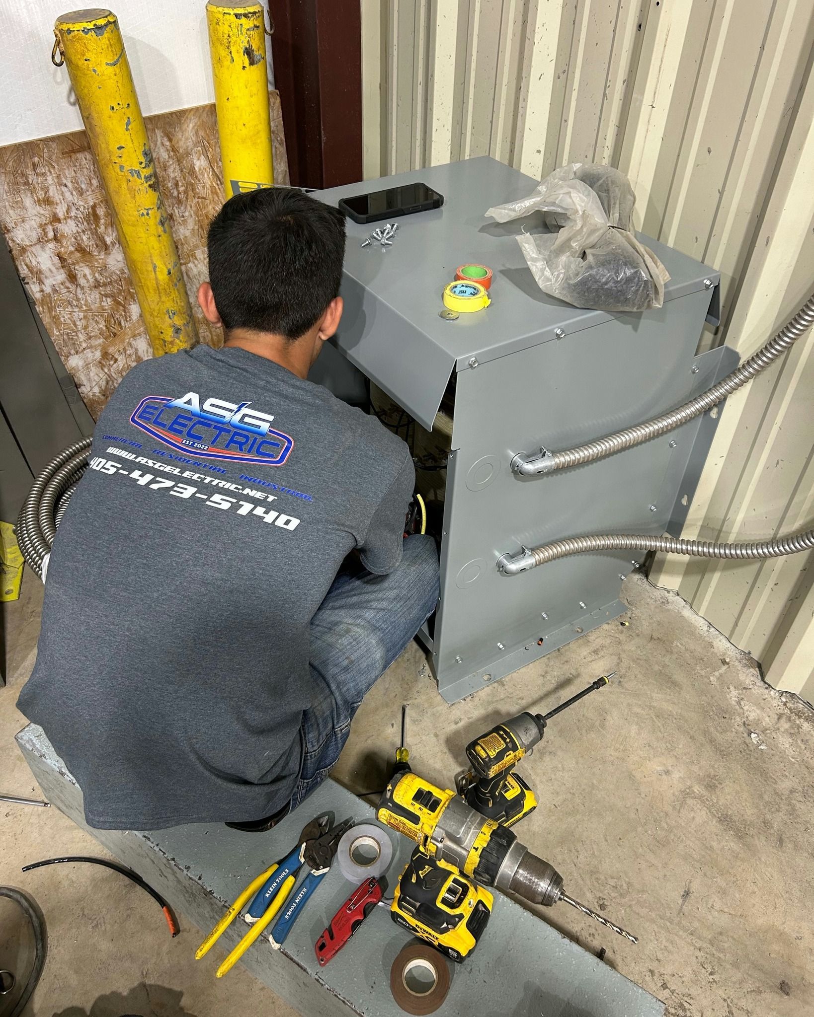 A person in a gray shirt working on electrical equipment, tools and cables visible.