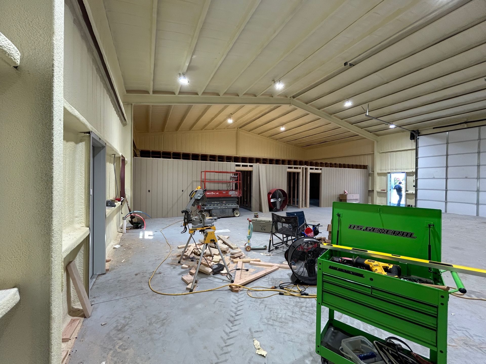Interior of a building under construction. Workers and tools are visible. Beige walls, gray floor, bright lights.