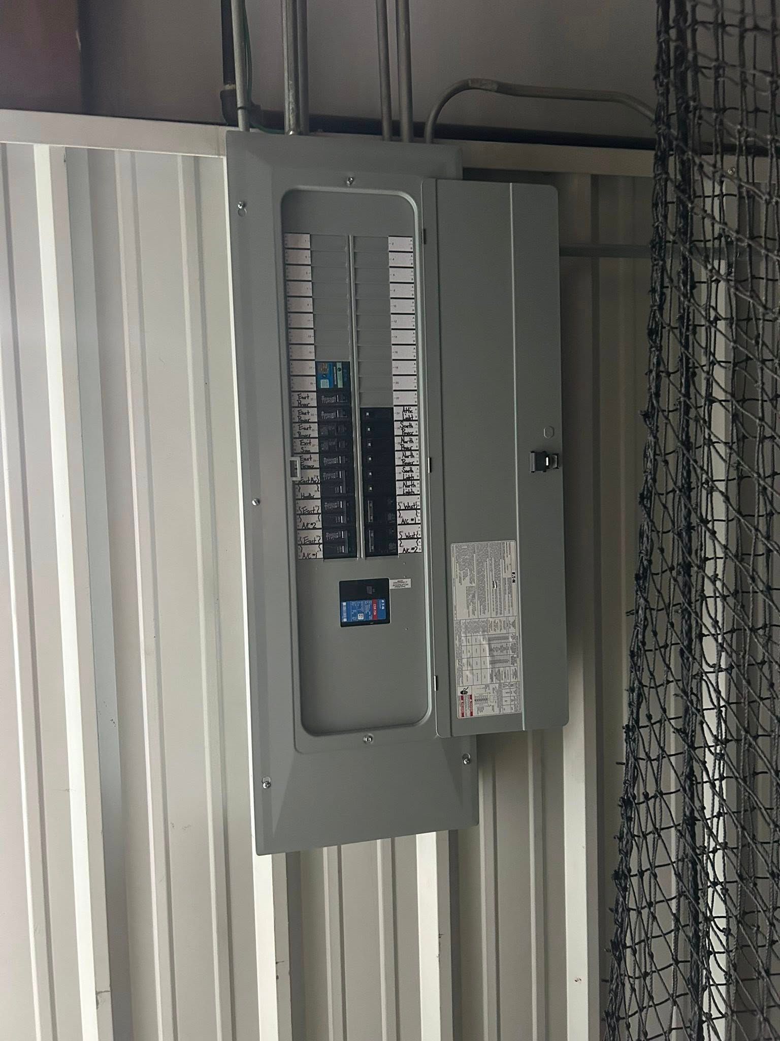 Gray electrical panel mounted on a corrugated metal wall, with some breakers flipped on.