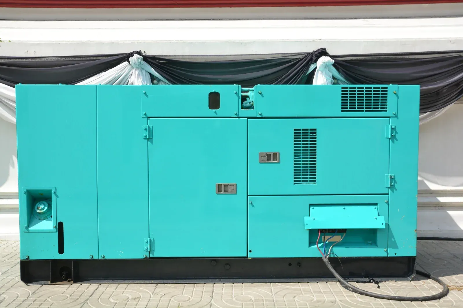 Turquoise industrial generator against a white wall with black draped fabric.