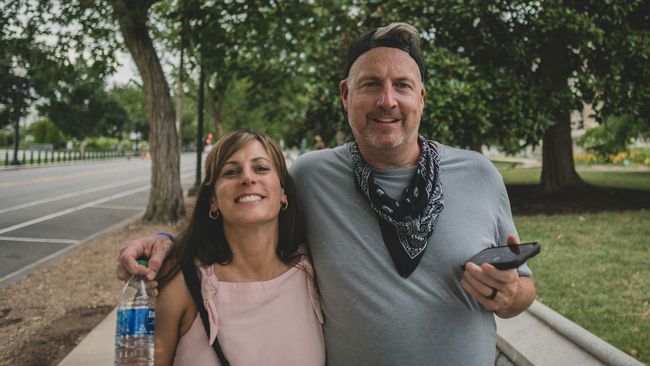 A man and a woman are posing for a picture in a park . the woman is holding a bottle of water.