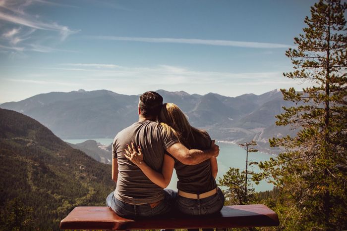 A man and a woman are sitting on a bench overlooking a lake and mountains.