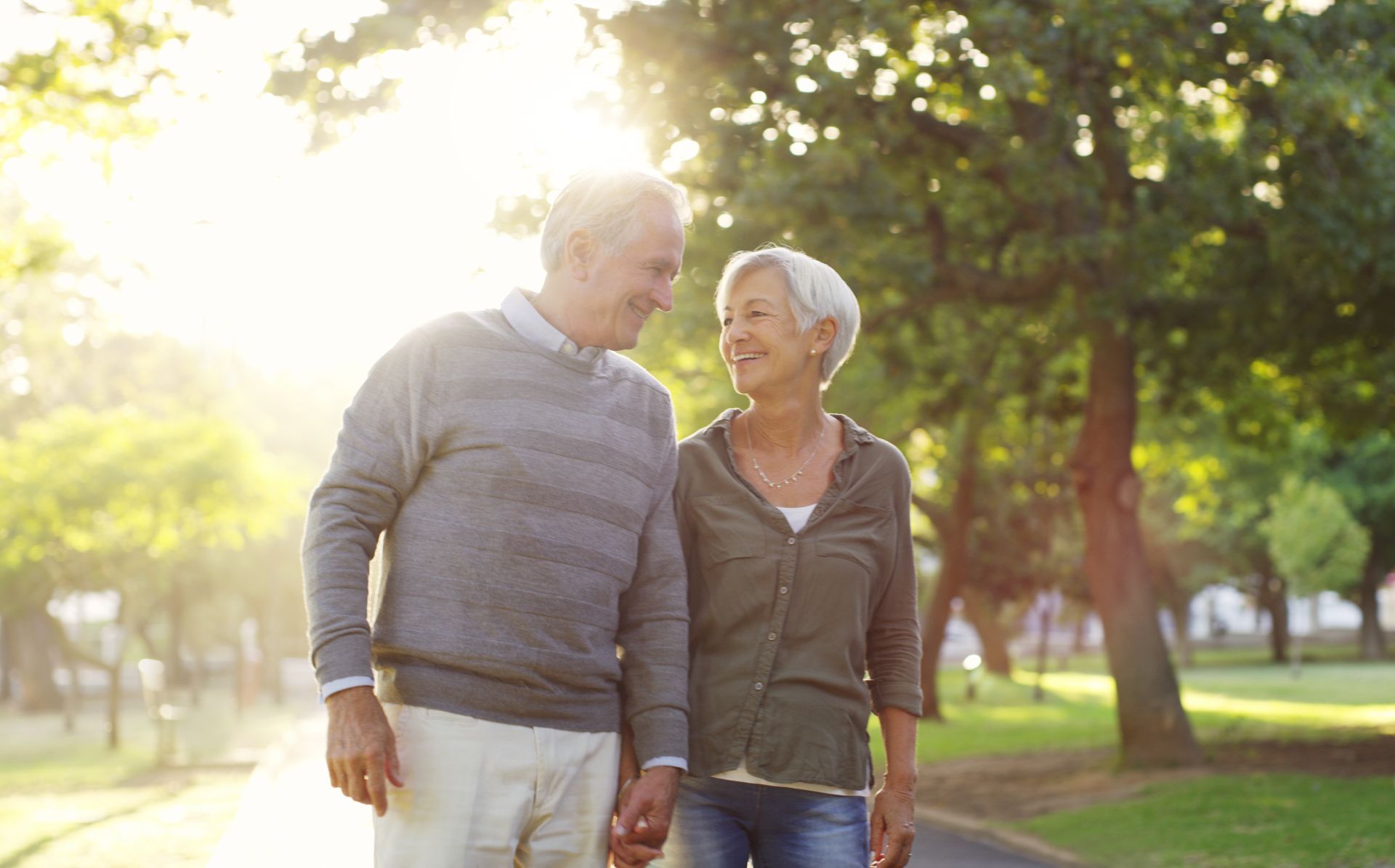 An elderly couple is walking down a path in a park holding hands.