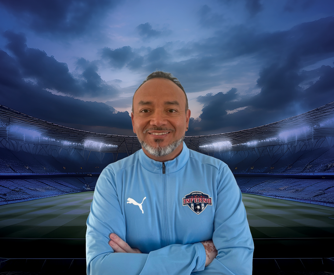 Coach Paul Tafur in light blue jacket smiling, in front of a soccer stadium background.