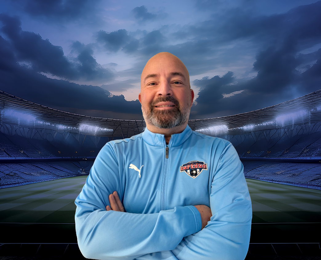Coach Piere Garcia with crossed arms wearing a light blue jacket with a soccer team logo; stadium background.