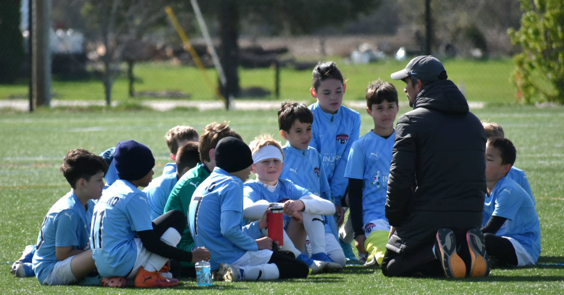 Youth soccer team huddles on a green field, listening to coach.