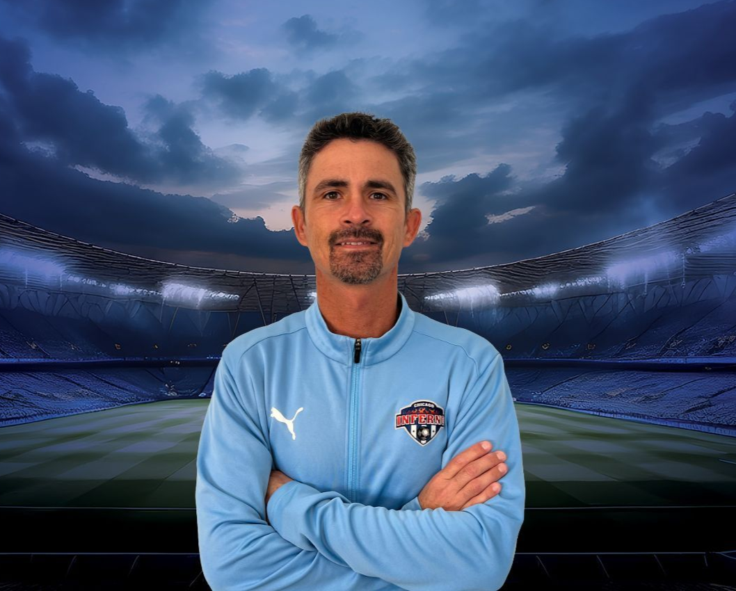 Coach Ewerton Andrade in light blue jacket with arms crossed, in a soccer stadium, cloudy sky background.