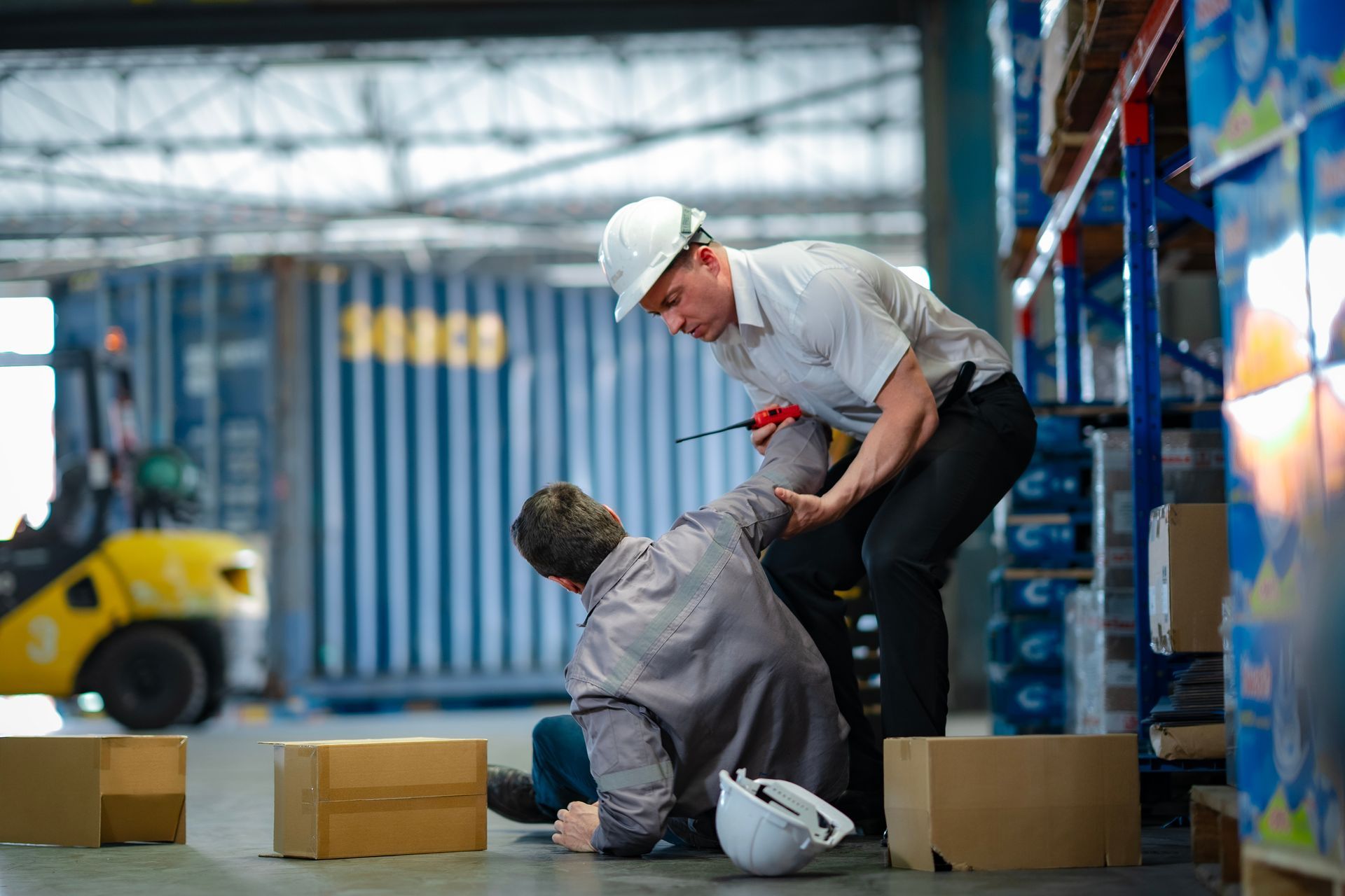 Warehouse worker helping injured colleague after a fall. Boxes and forklift visible.