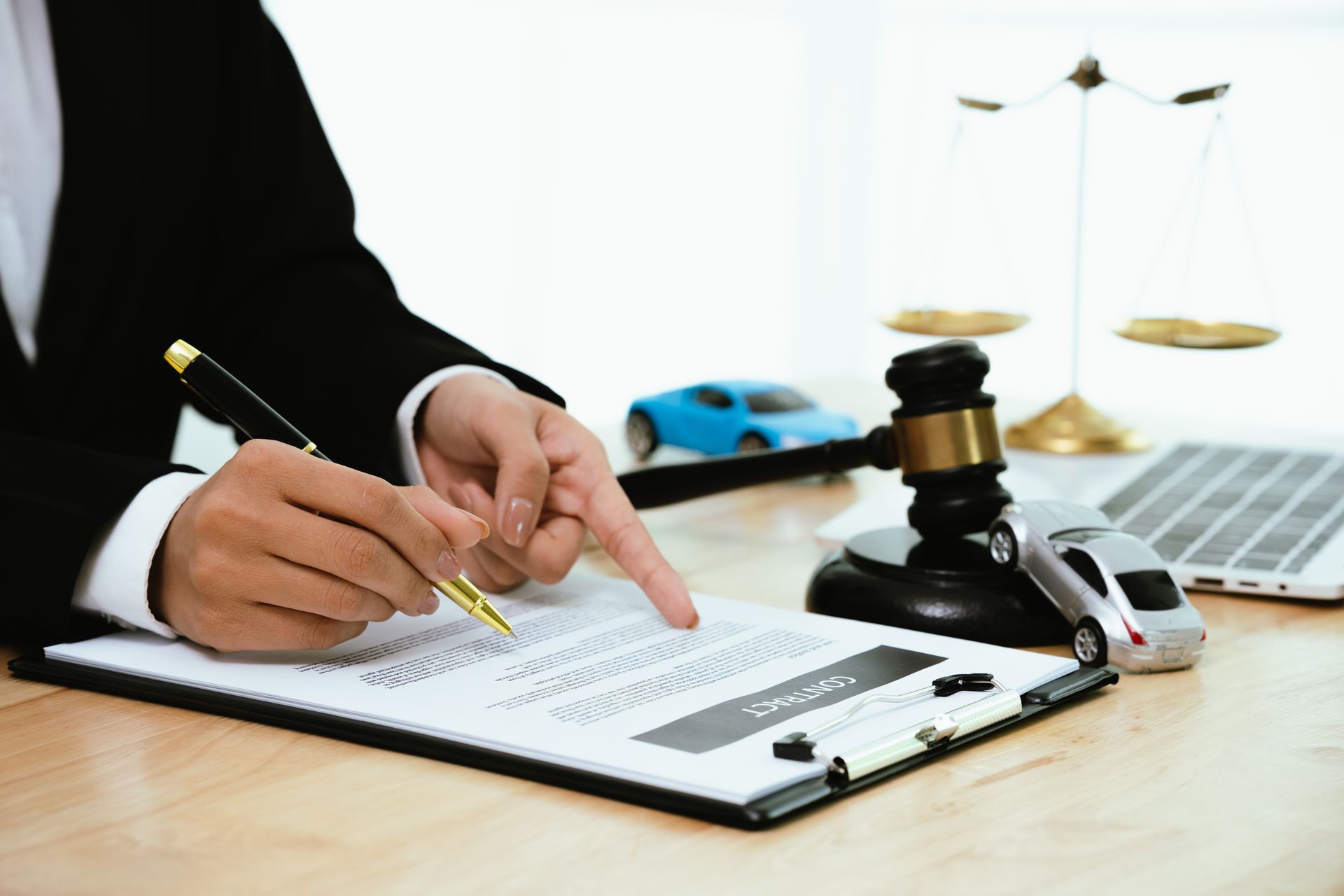 Person in suit pointing at contract on desk with toy cars, scales, and gavel.