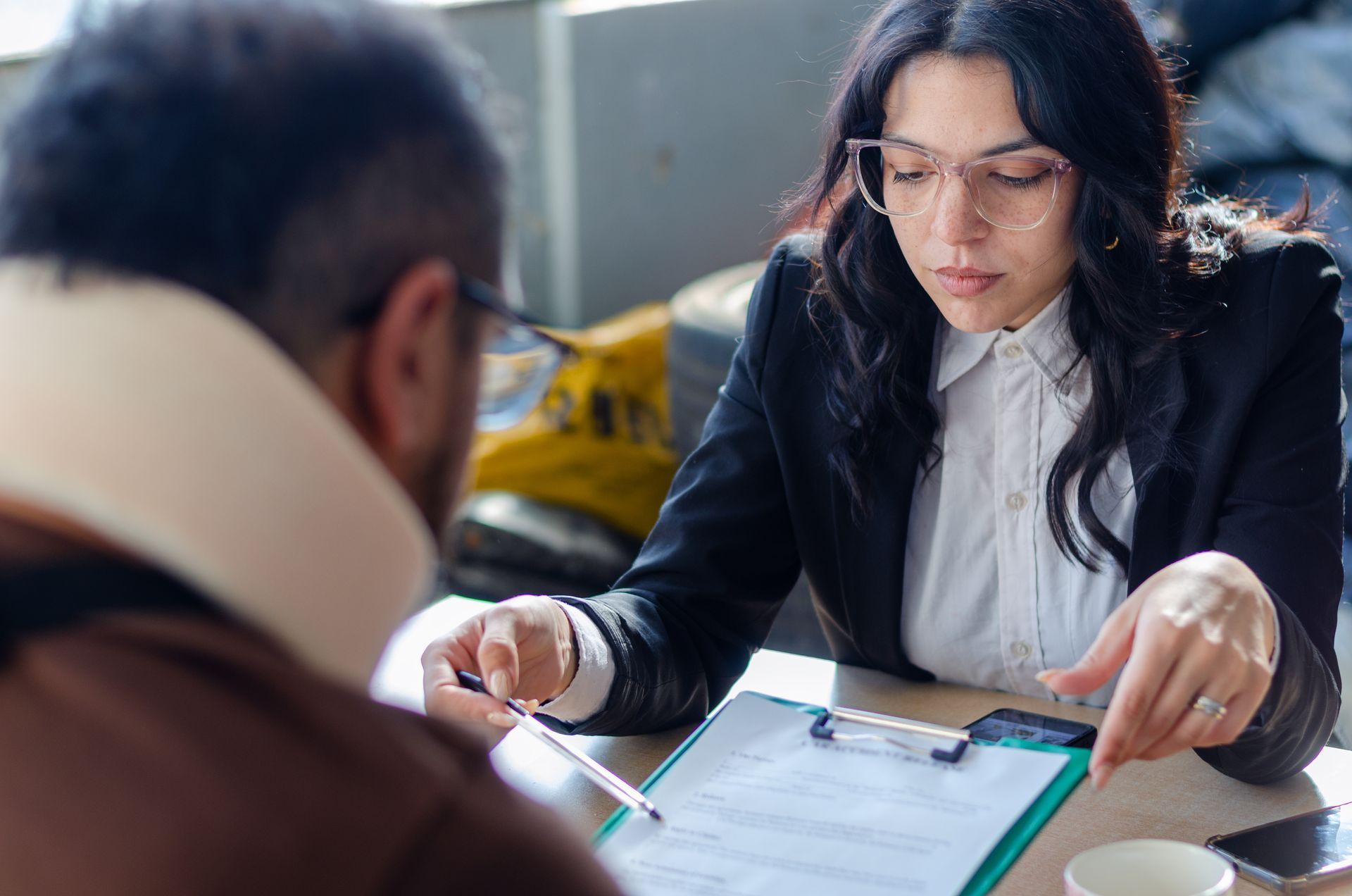 Woman in glasses points at paperwork with a man wearing a neck brace, at a table.