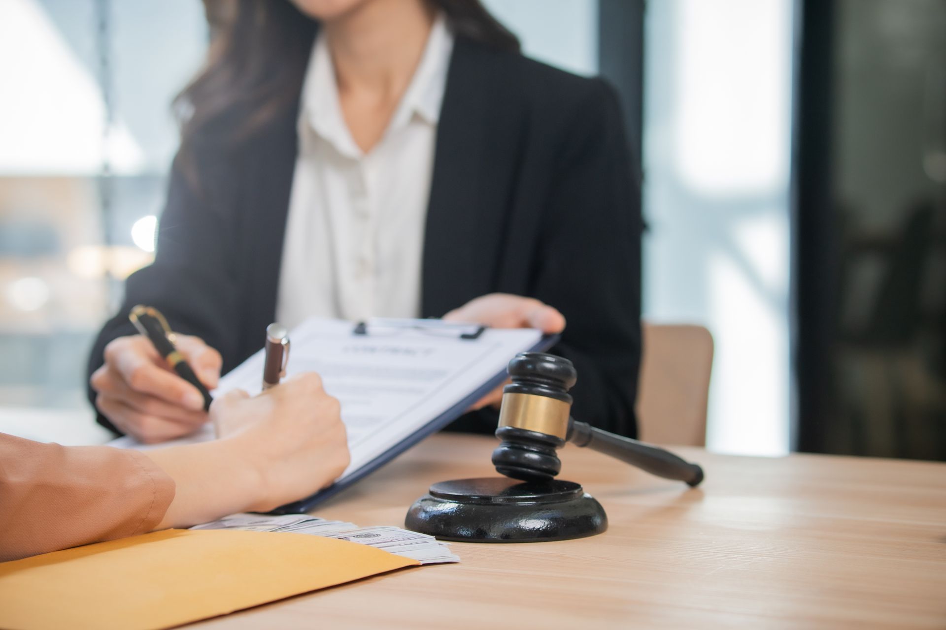 Person signing document; gavel on desk; another person holds clipboard.