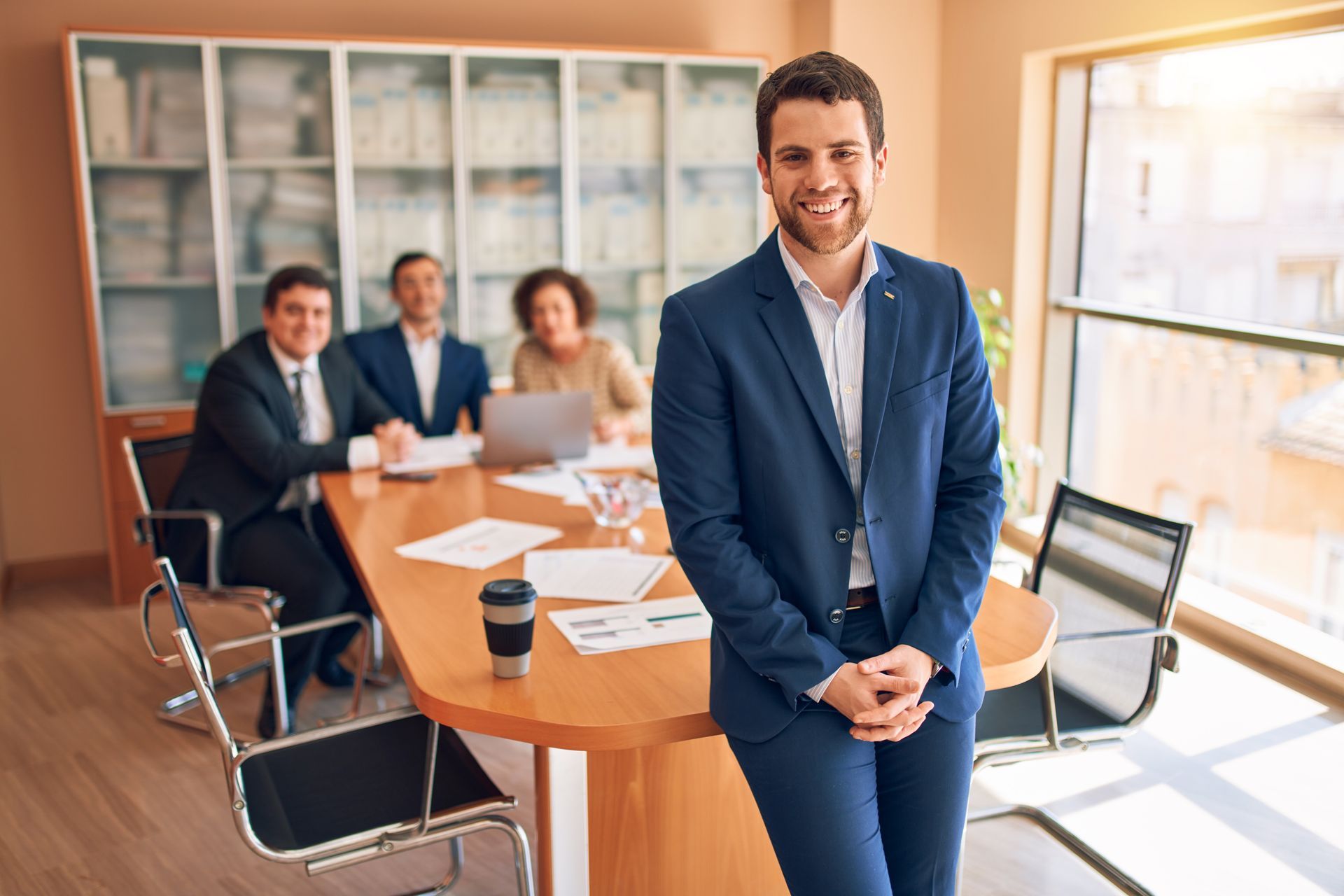 Man in blue suit smiling, leaning on a table in an office, with coworkers at a conference table. Man in blue suit smiling, leaning on a table in an office, with coworkers at a conference table.