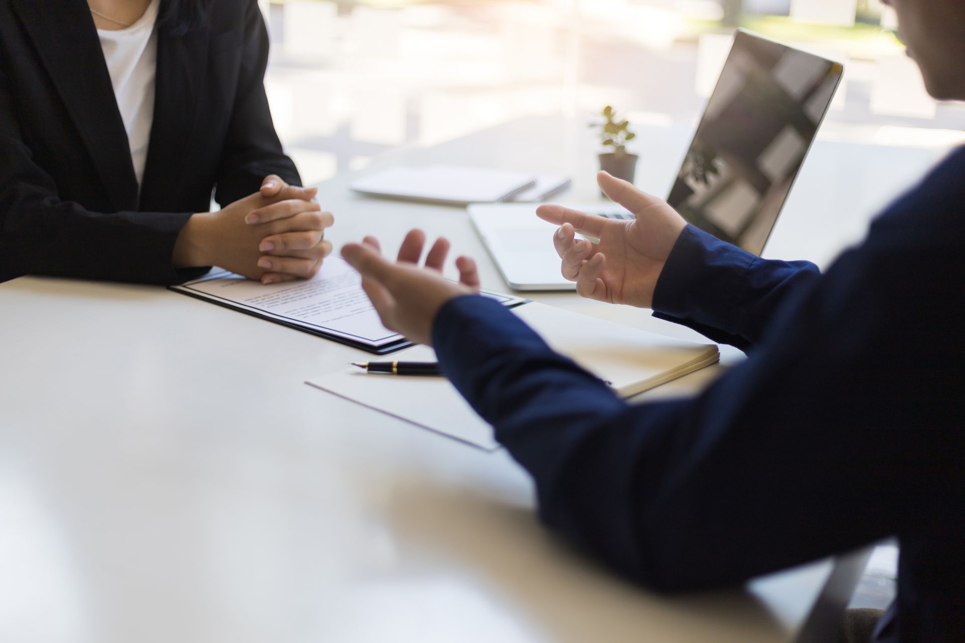 Two people in business attire at a table; one speaking, gesturing; the other listening, hands clasped.