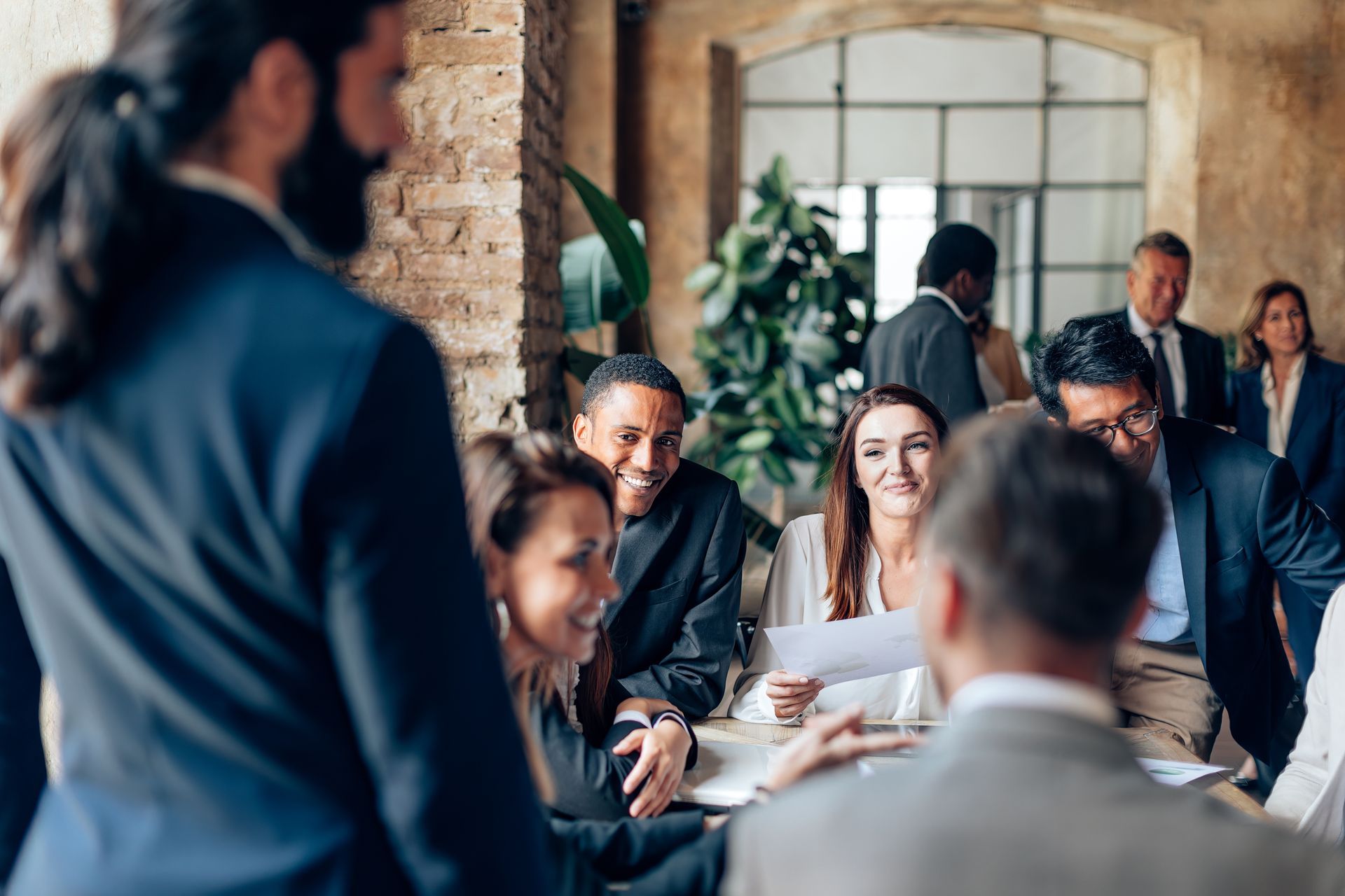 Businesspeople in a meeting, some seated at a table, others standing. Smiling faces, formal attire, in an office space. Businesspeople in a meeting, some seated at a table, others standing. Smiling faces, formal attire, in an office space.