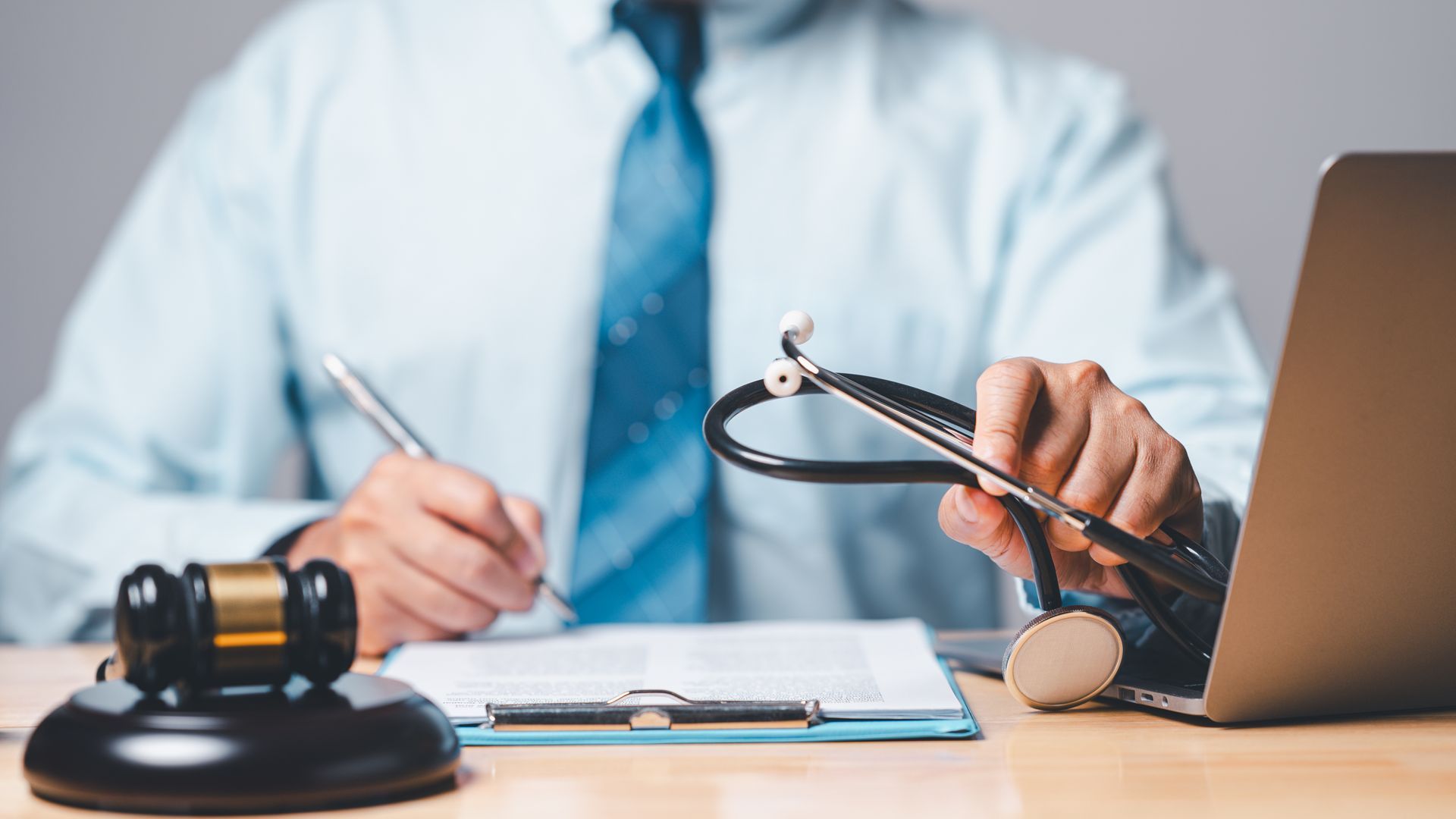 A person holding a stethoscope, gavel, and laptop on a desk, possibly a medical legal setting.