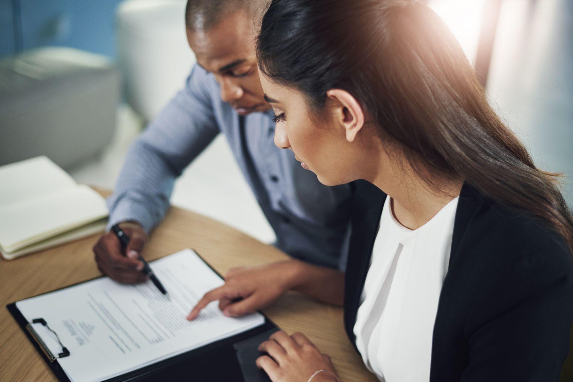 Two people reviewing documents at a desk; one points while the other looks on. Two people reviewing documents at a desk; one points while the other looks on.