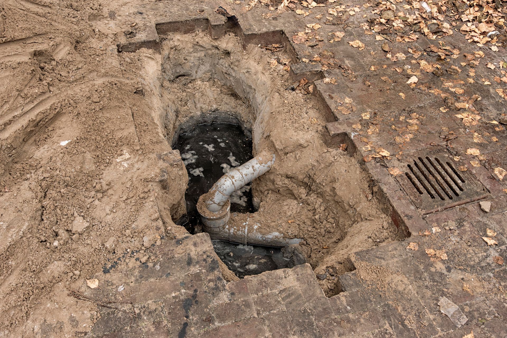 Open trench revealing a pipe, near a drainage grate, set in dirt.