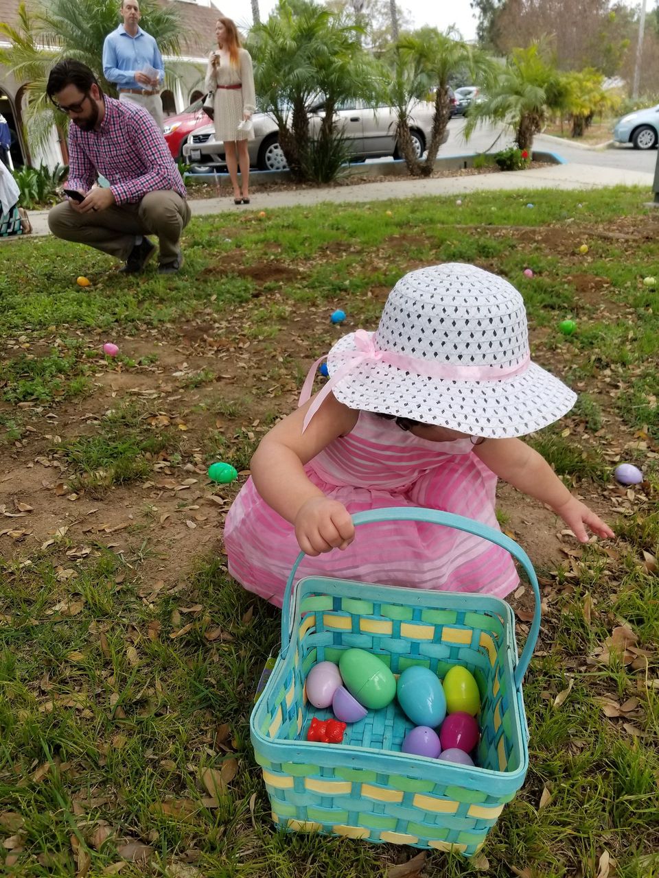 A little girl is sitting in the grass next to a basket of easter eggs.