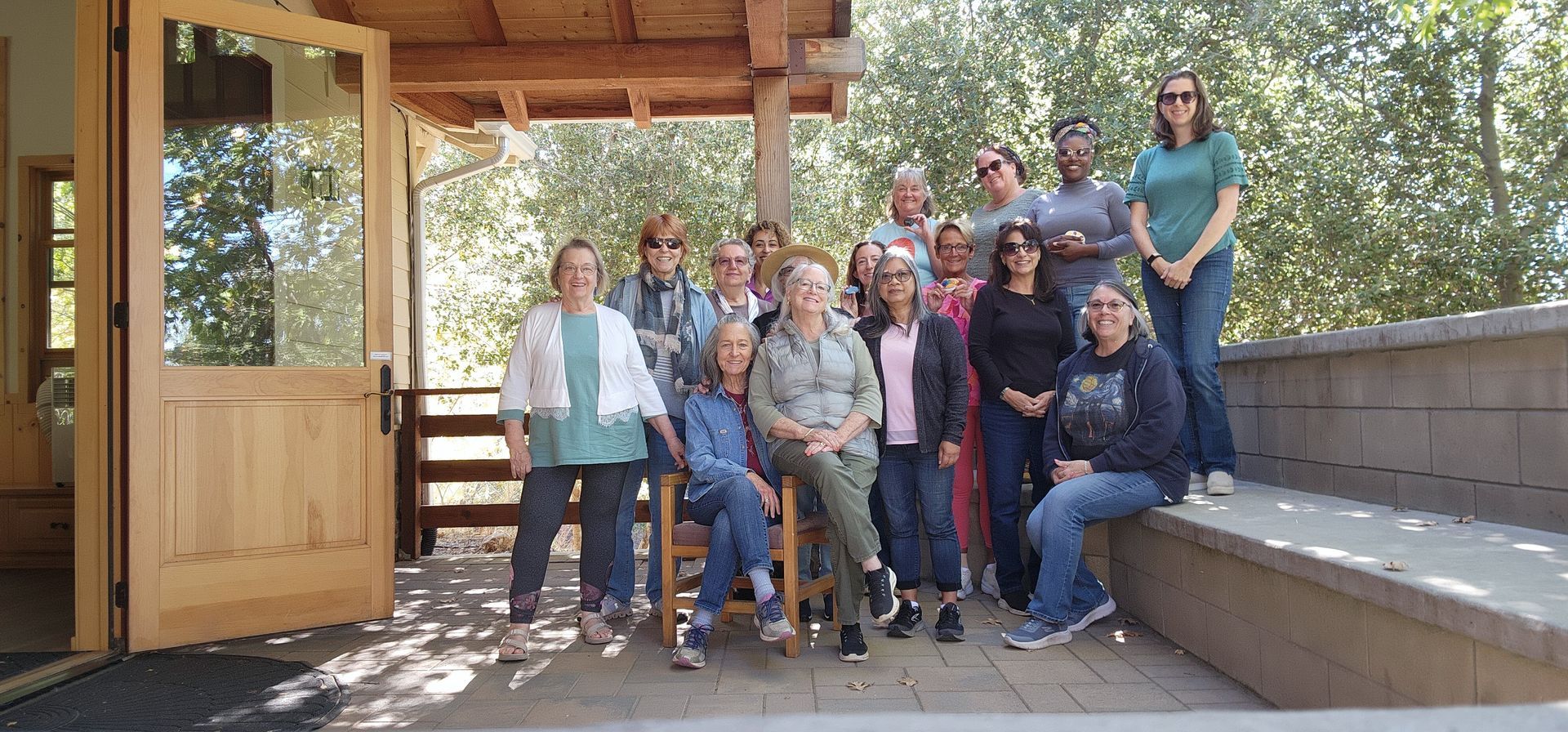 Group of people standing on a porch; some are smiling. The setting is outdoors, with wooden accents.