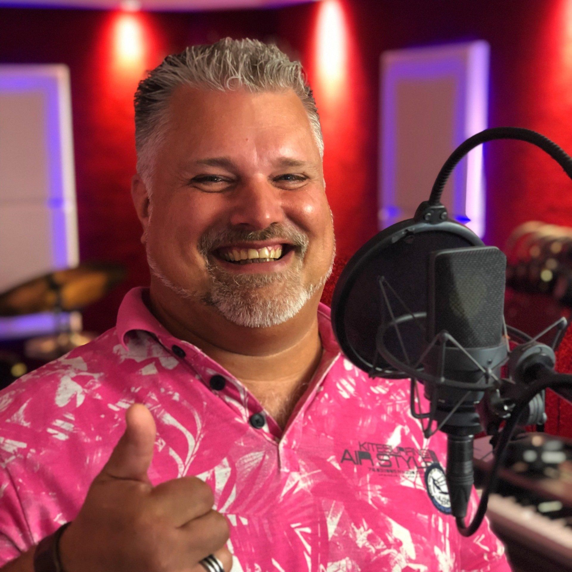 Een man in een roze shirt met een microfoon in een opnamestudio steekt zijn duim omhoog.