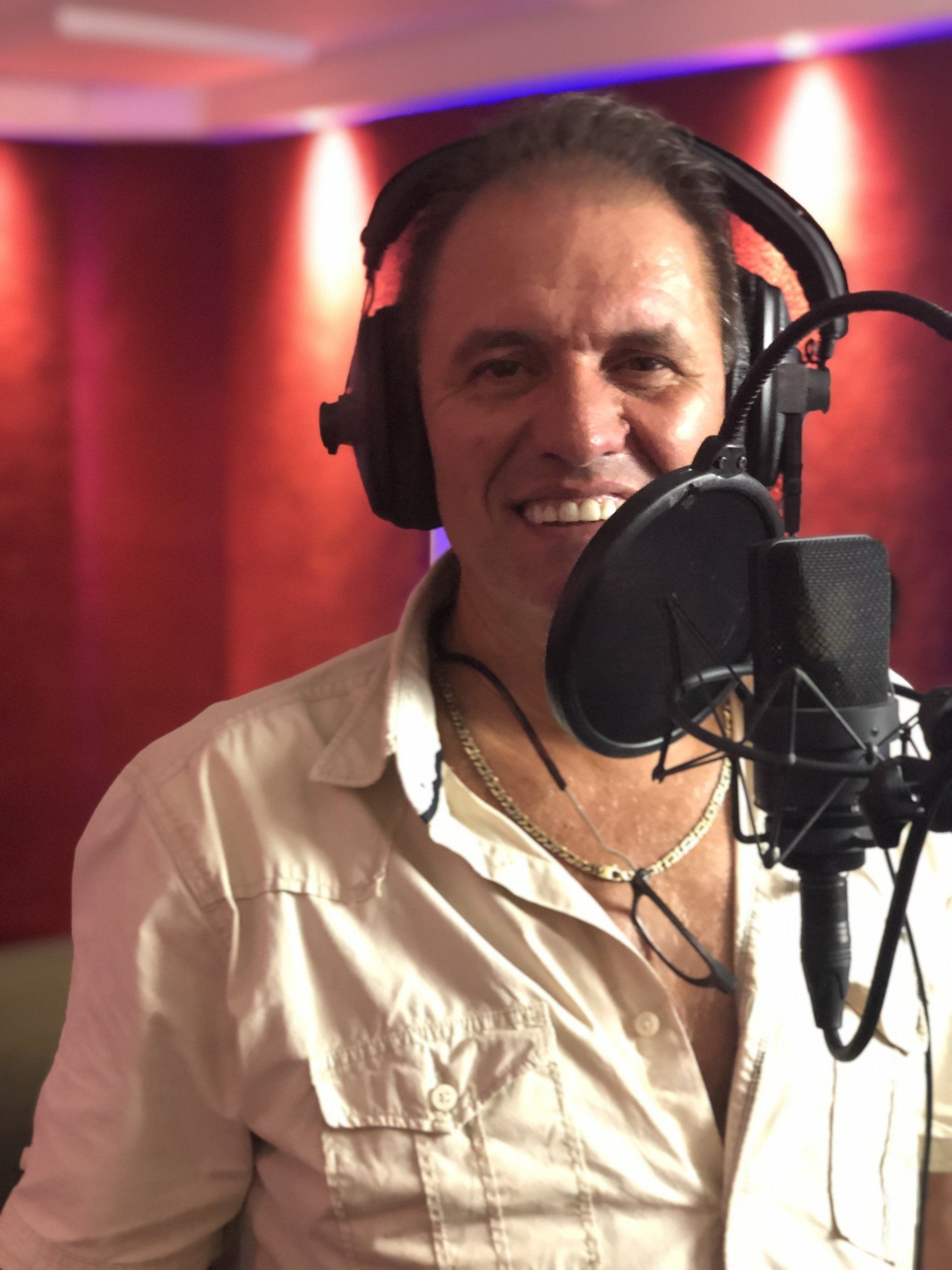 Man in opnamestudio met koptelefoon, lachend naar de microfoon. Rode achtergrond.