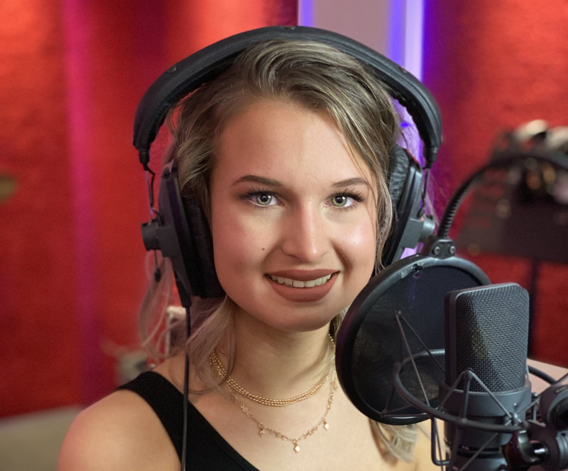 Vrouw met koptelefoon in een opnamestudio, lachend. Ze kijkt in de camera, met een microfoon voor zich. Rode achtergrond.