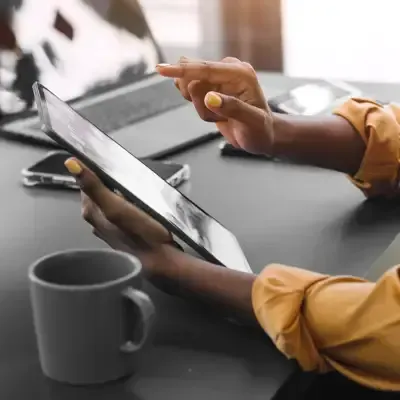 Hands using a tablet at a desk with a coffee mug and laptop nearby