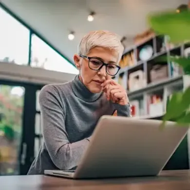 Person in glasses working on a laptop in a bright café, hand resting on chin.