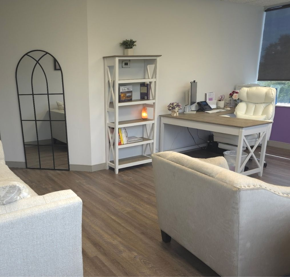 Cozy office with desk, bookshelf, mirror, and plush chairs and couch in a bright room with wood floors