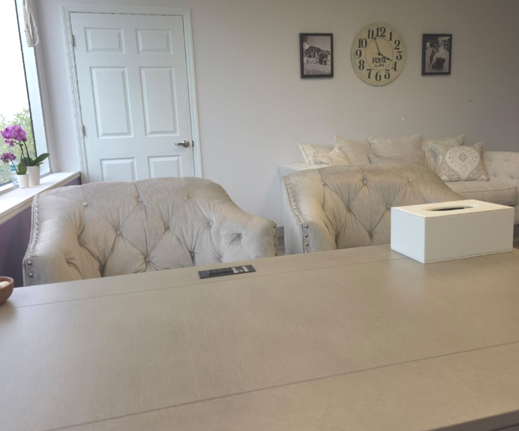 Bright living room with beige sofa, tufted chair, wall clock, and large table in the foreground.