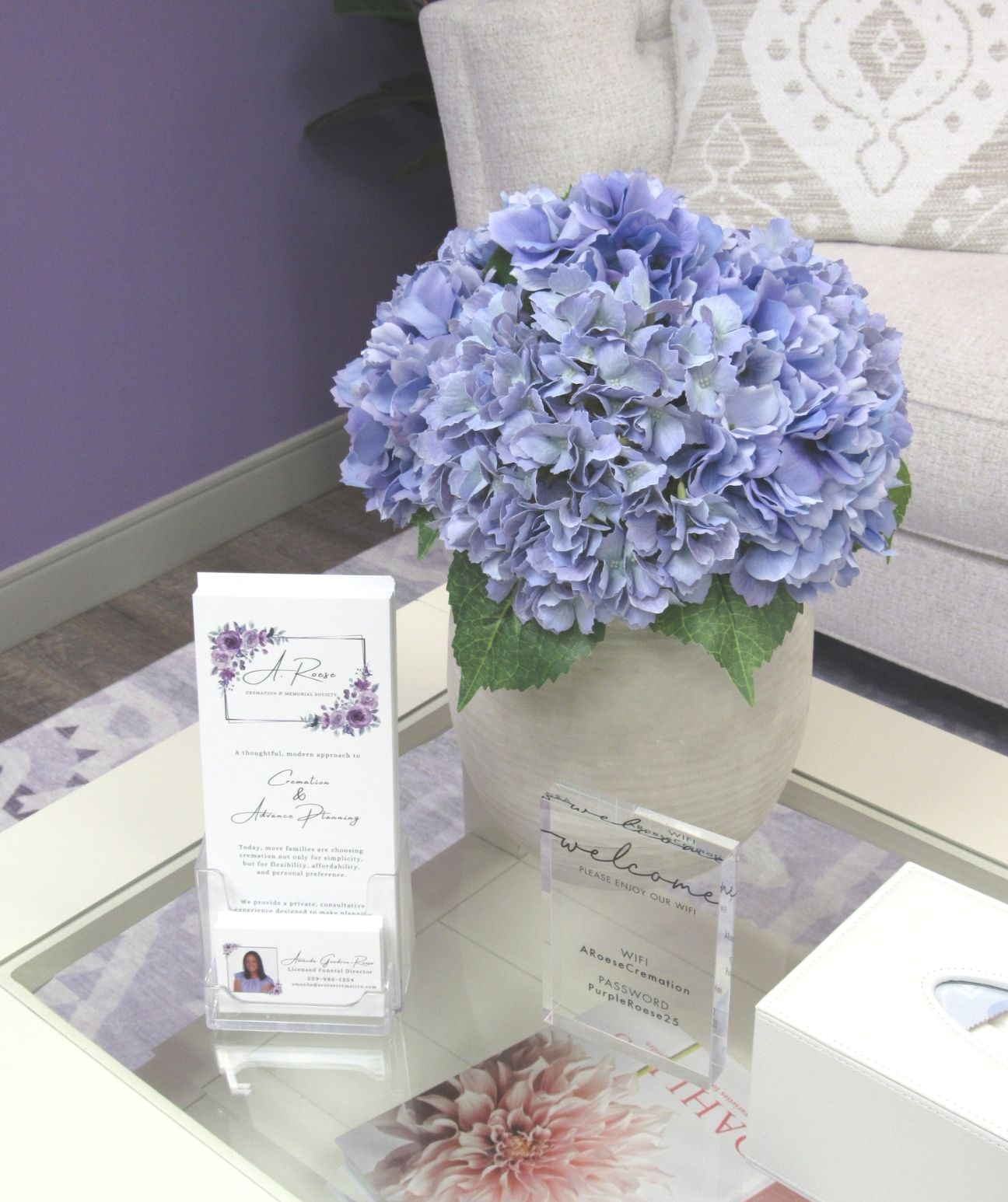 Blue hydrangea bouquet in a vase on a glass table with decorative cards and a sofa in the background