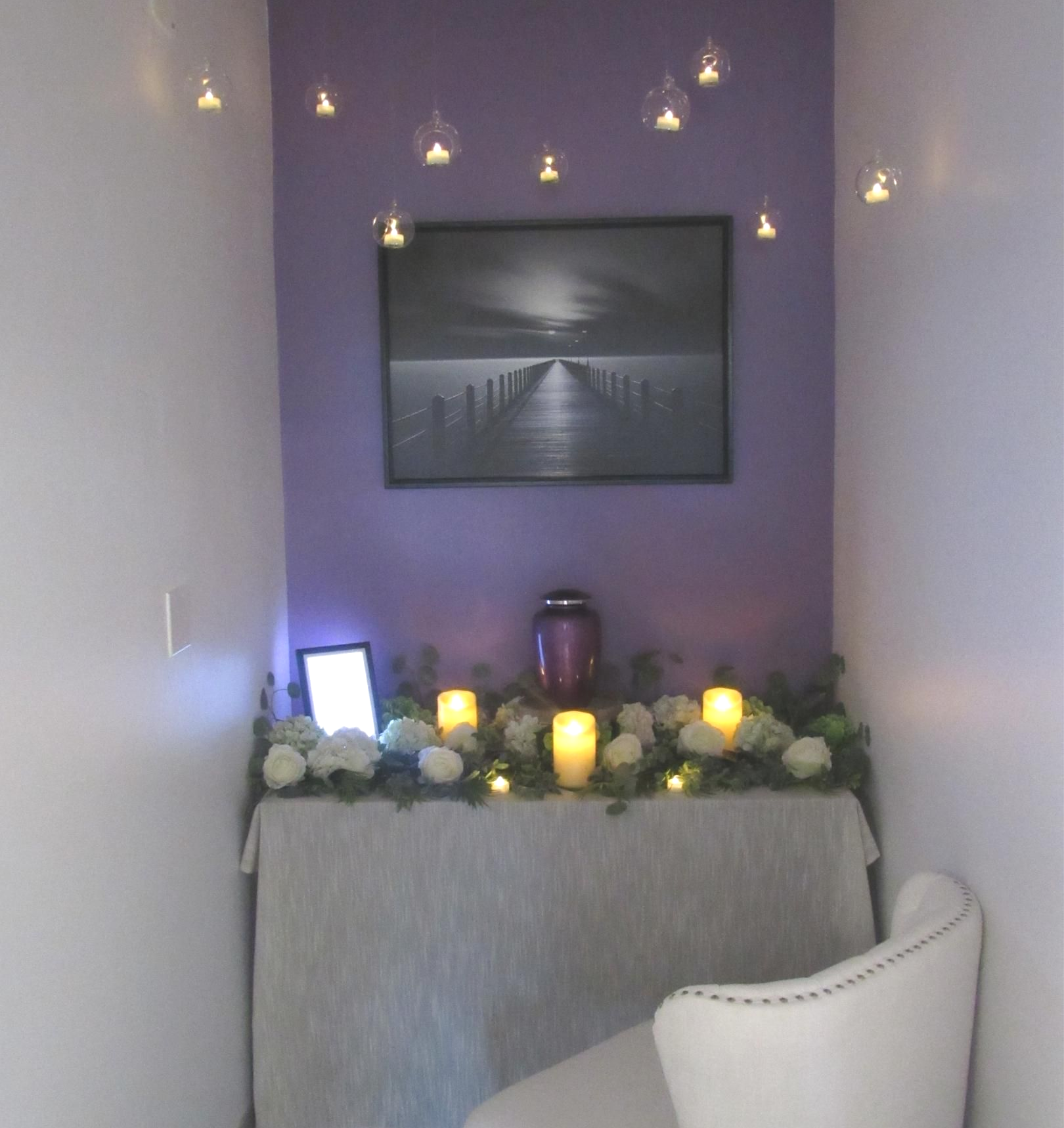 Dim cozy alcove with candles, fairy lights, a framed picture, and a white chair.