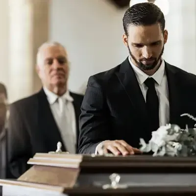 Man in black suit signing a document at a funeral, with an older man standing behind him.