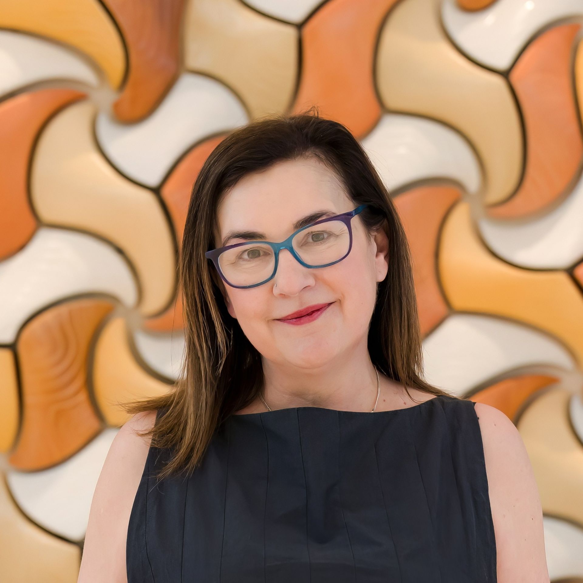 Aurora Bariatrics team member Justine Giles in a sleeveless navy dress with glasses, smiling in front of an orange and cream geometric wall