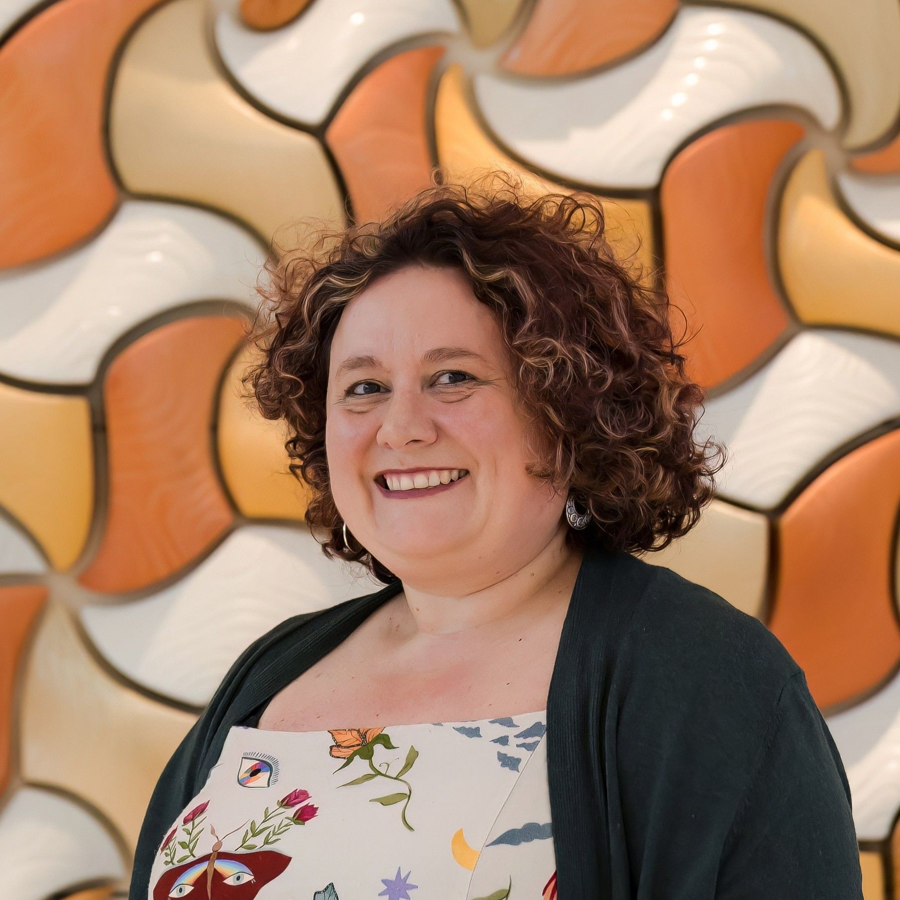 Aurora Bariatrics team member Claire Brown with curly hair wearing a patterned dress and dark cardigan, smiling in front of a geometric orange and cream background