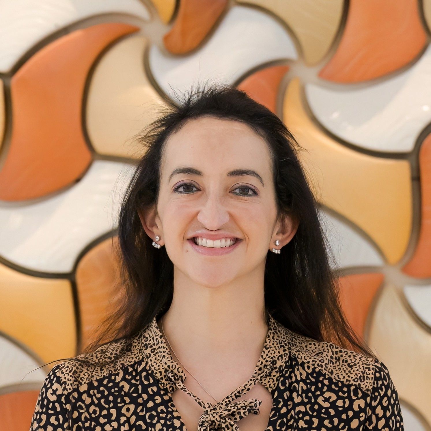 Aurora Bariatrics team member Dr Aimee Pikoos with long dark hair, wearing a leopard print blouse and earrings, smiling in front of a warm-toned mosaic tile wall