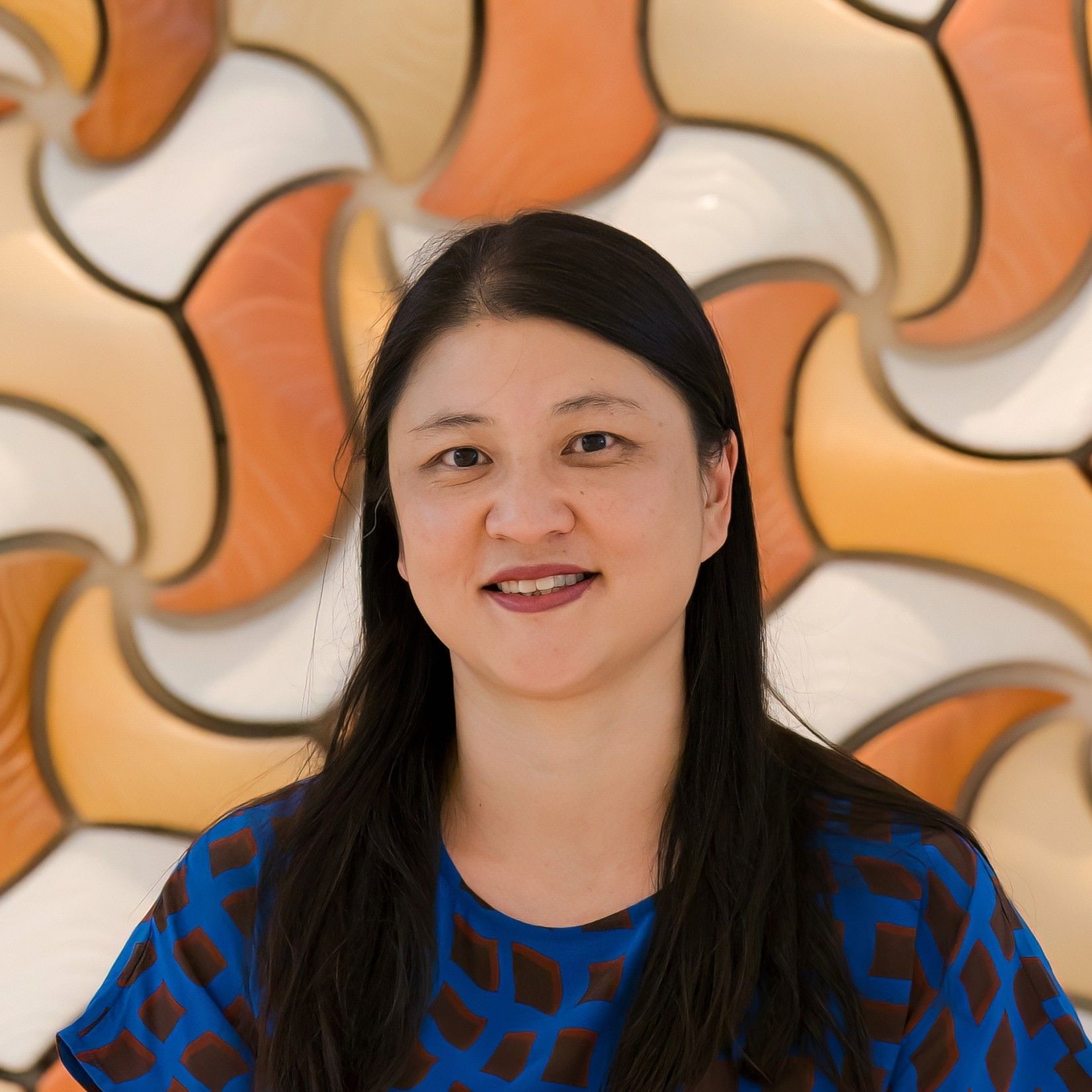 Aurora Bariatrics team member Dr Cindy Ng with long dark hair wearing a blue patterned top, smiling in front of a curved orange and cream tiled background