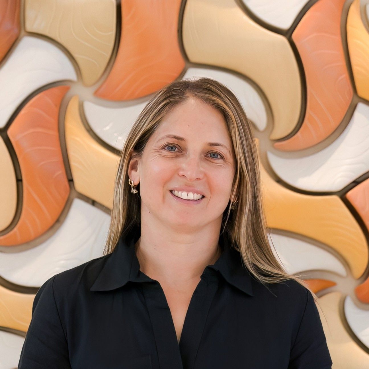 Aurora Bariatrics team member Loren Muhlmann with long straight blonde hair wearing a black blouse, smiling in front of a curved orange and cream mosaic tile wall