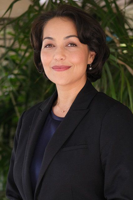 A woman in a black jacket and blue shirt is standing in front of a plant.